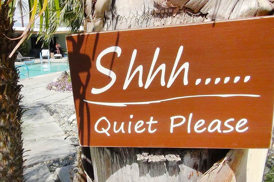 Wooden 'Shhh... Quiet Please' sign at poolside with palm trees and pool visible