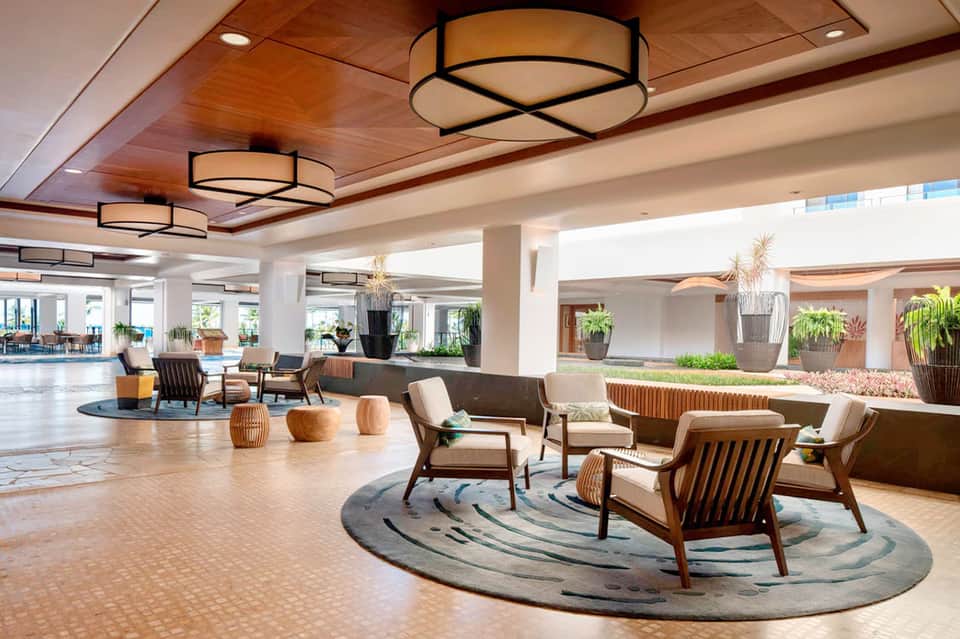 Spacious hotel lobby with wooden ceiling, mid-century furniture, and potted plants throughout