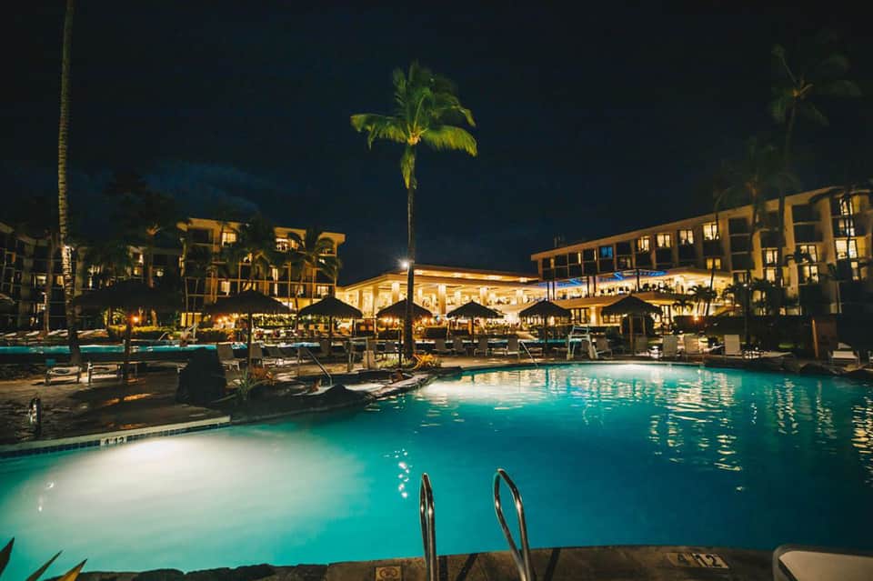 Resort pool illuminated at night with palm trees and hotel buildings