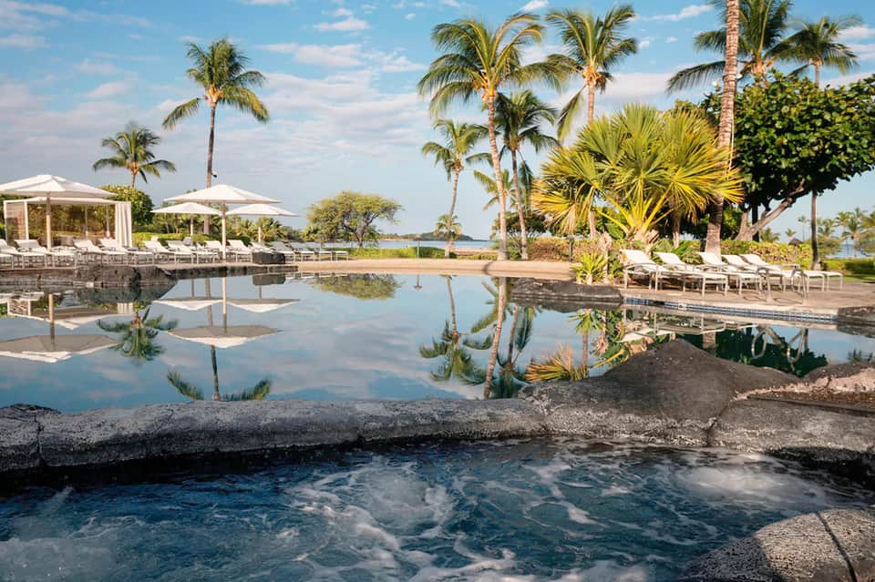 Resort pool with white umbrellas, loungers, and palm trees reflecting in calm water