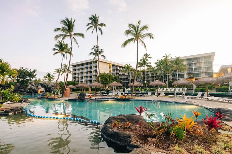 Tropical resort pool with thatched cabanas, palm trees, and multi-story hotel buildings