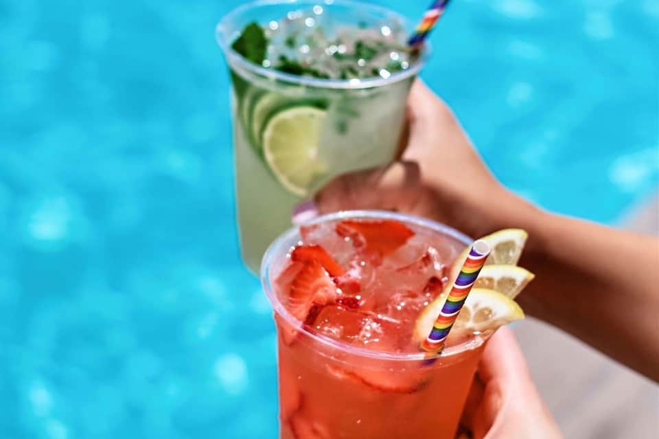 Colorful cocktails held by hands poolside with tropical blue water in background