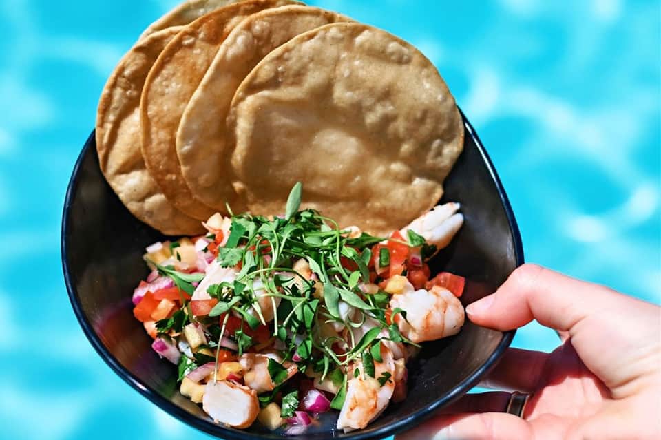 Hand holding black plate with shrimp and vegetable appetizer with pita bread against bright blue sky background.