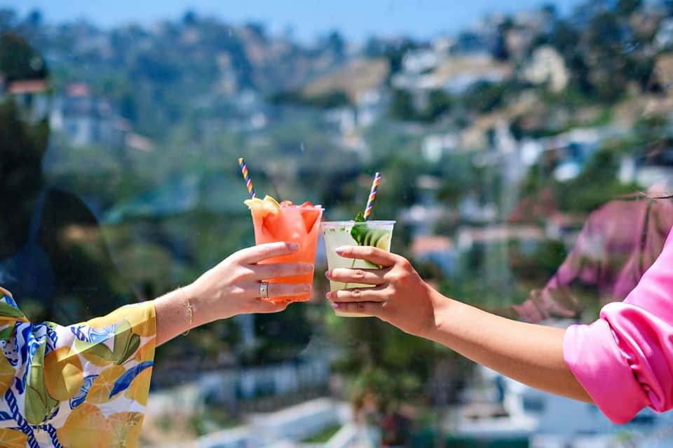 Two hands toasting colorful cocktails overlooking a blurred hillside village landscape