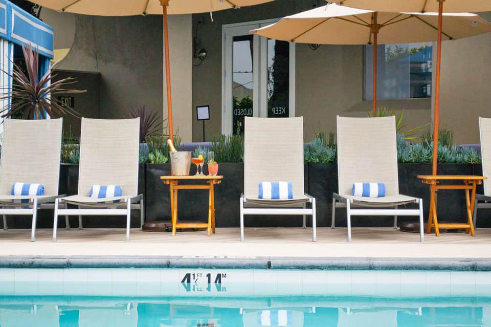 Poolside lounge chairs with blue and white striped cushions under tan umbrellas beside a swimming pool