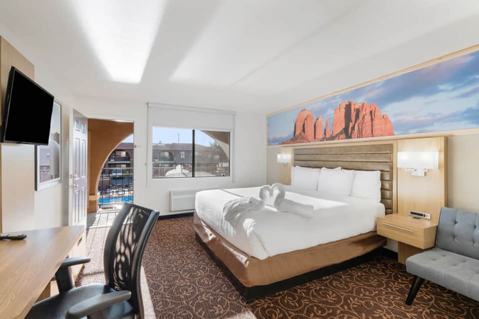 Modern hotel bedroom with queen bed, work desk, mountain artwork, and balcony with city views