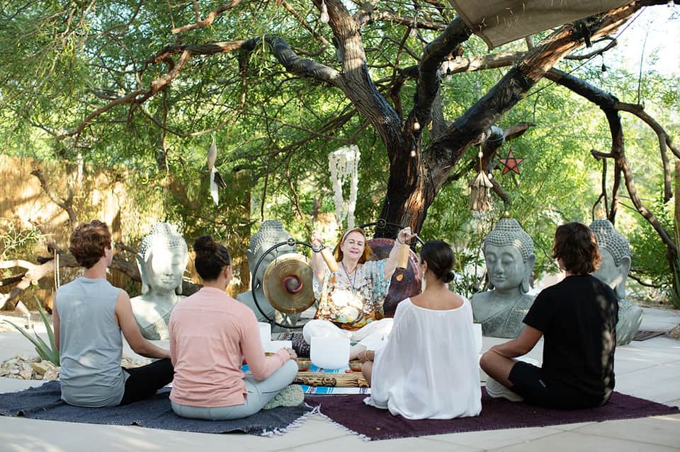 Group meditation circle under large tree with Buddha statues and gong at wellness retreat