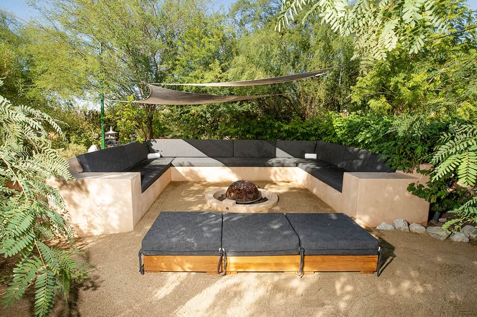 Backyard fire pit lounge area with built-in stone seating, shade sail, lush green trees
