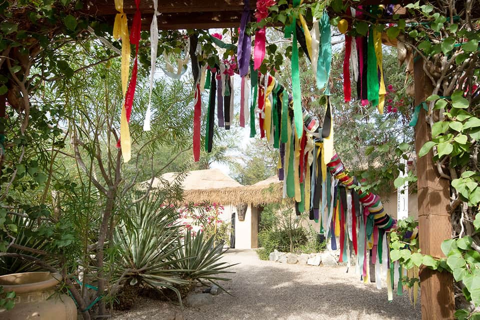 Tropical pathway with colorful fabric ribbons overhead, lush vegetation, and thatched structures