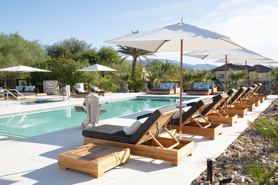 Desert resort pool with wooden lounge chairs, tan umbrellas, palm trees, and mountain views