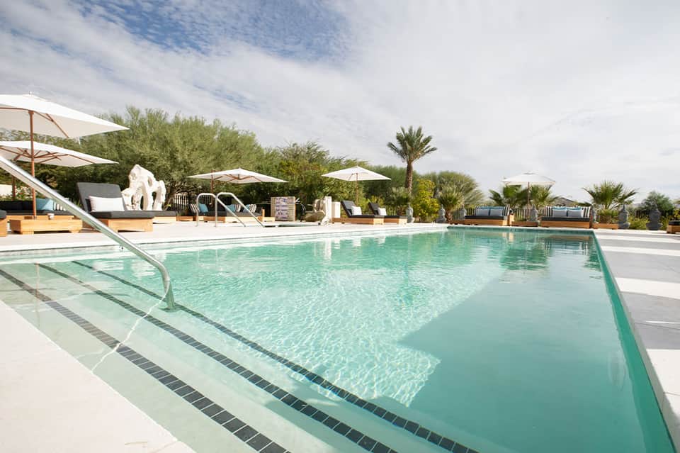 Resort swimming pool with turquoise water, white umbrellas, loungers, and palm trees in desert setting