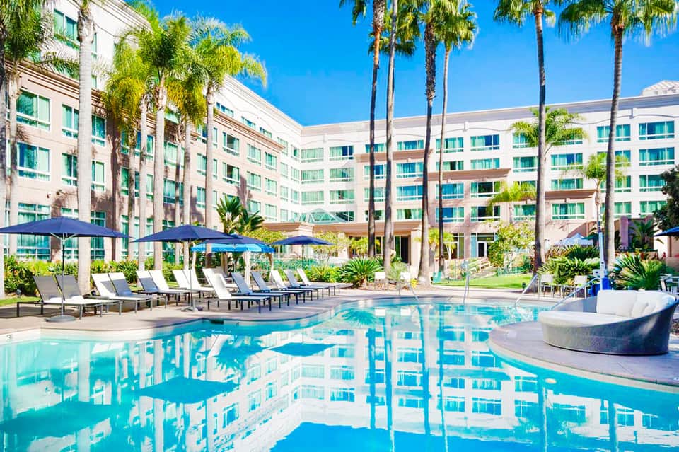 Resort courtyard with crystal blue pool, palm trees, lounge chairs, blue umbrellas, and multi-story hotel building