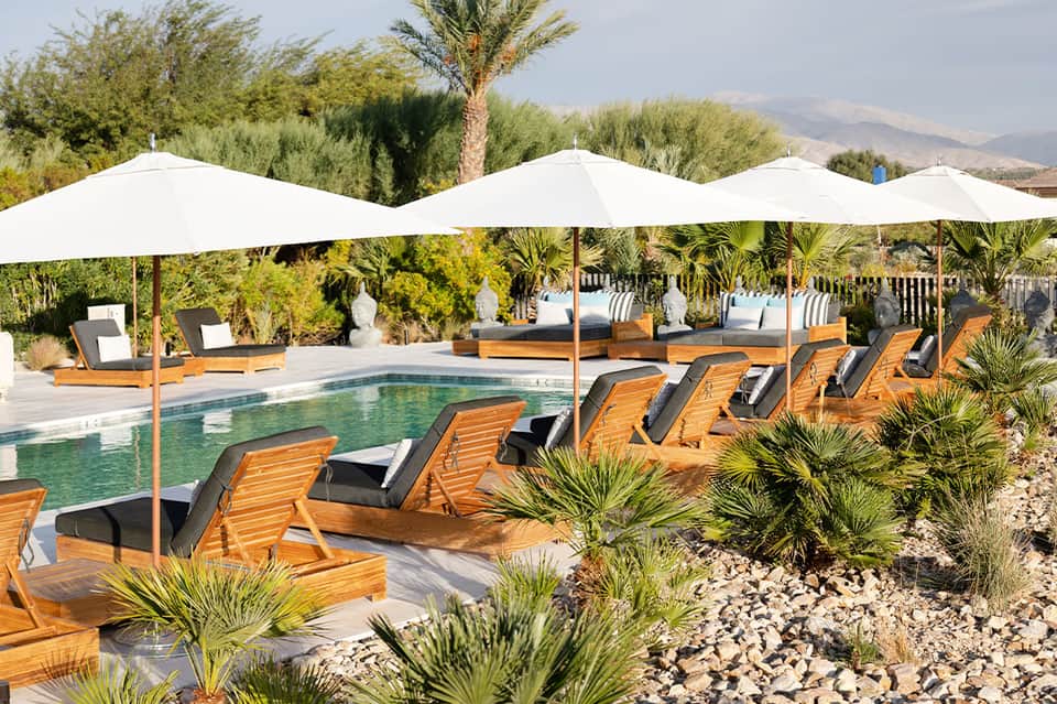 Luxury resort pool with white umbrellas, wooden loungers, mountain views, and desert landscaping