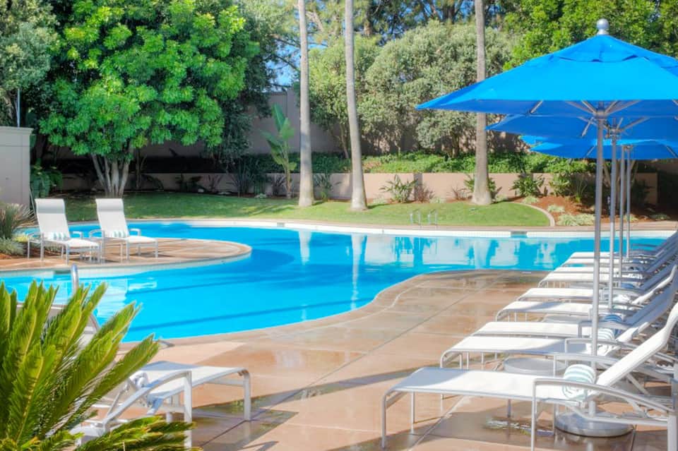 Curved blue resort pool with lounge chairs, blue umbrella, and palm trees in landscaped garden setting