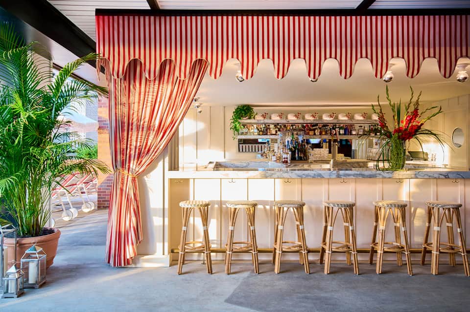 Beach bar with red and white striped awning, white bar counter, wicker stools, and tropical plants