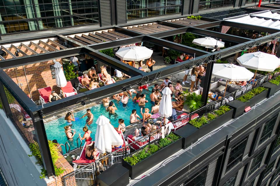 Aerial view of busy resort rooftop pool with swimmers, white umbrellas, and surrounding urban buildings