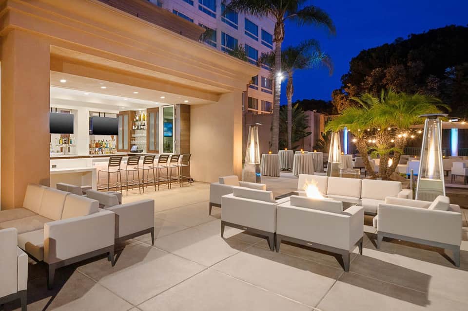 Evening outdoor bar and lounge area with white seating, illuminated bar counter, and palm trees under blue sky
