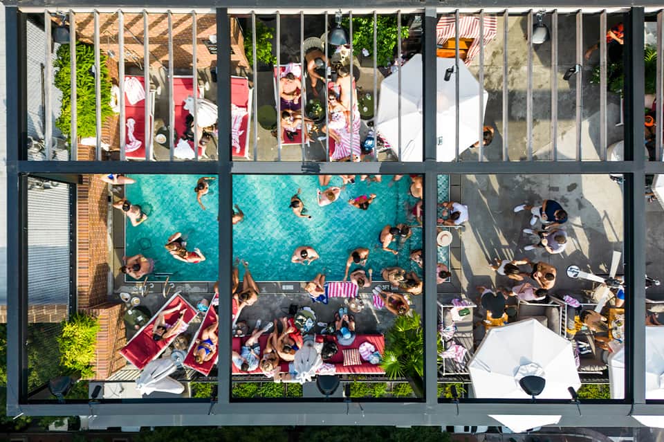 Aerial view of rooftop pool and terrace with guests, loungers, umbrellas, and surrounding cityscape