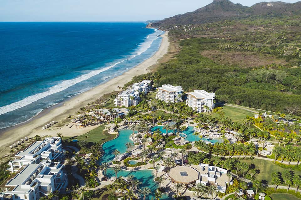 Aerial view of beachfront resort with pools, palm trees, and white buildings beside turquoise ocean