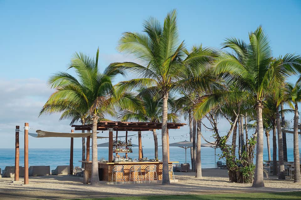 Beachfront bar with palm trees, wooden pergola, and ocean view at sunset
