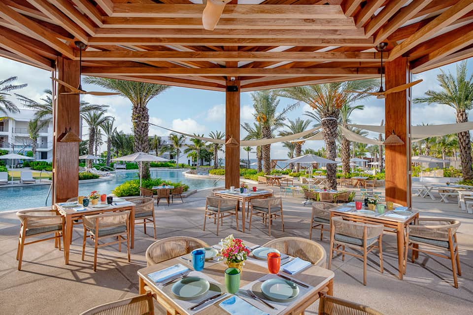 Upscale outdoor dining area with wooden beams, tables, pool views, and palm trees