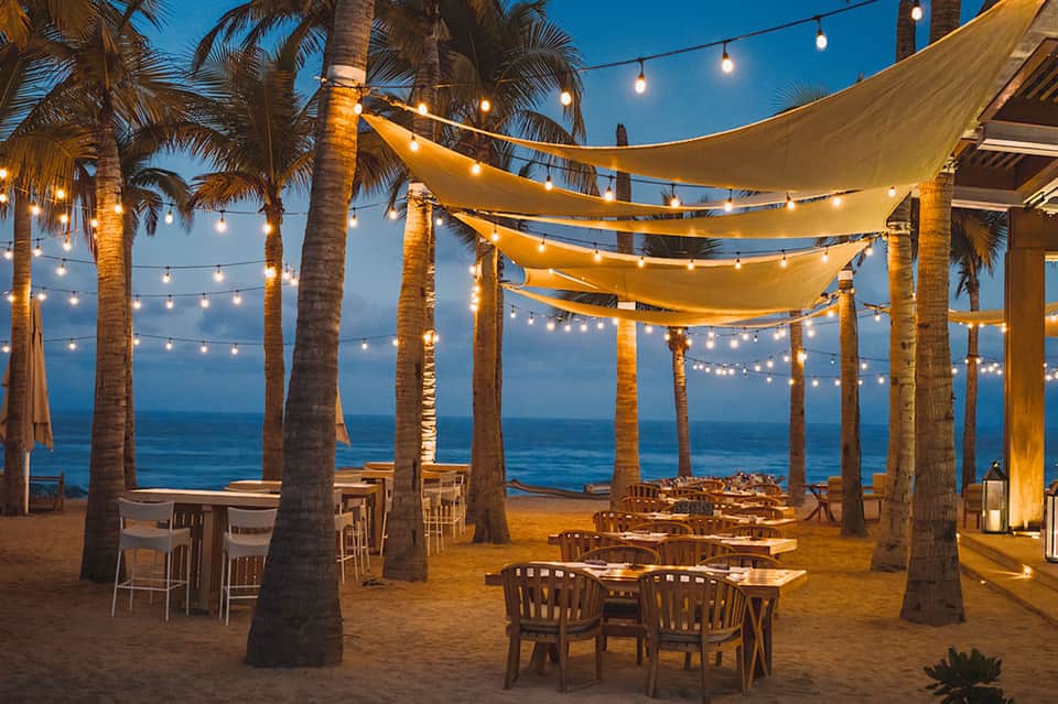 Beachfront dining area with palm trees, string lights, and ocean views at dusk