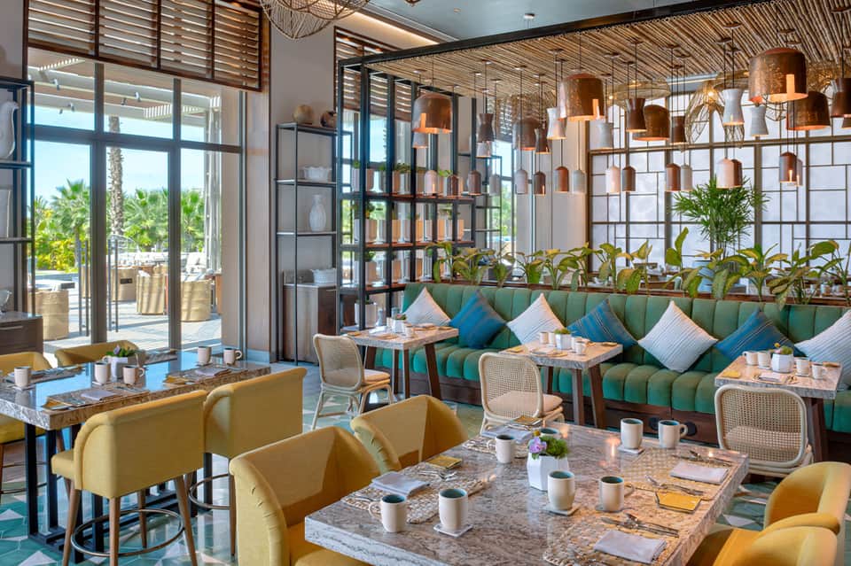Bright restaurant interior with green upholstered banquettes, copper pendant lights, and ocean views