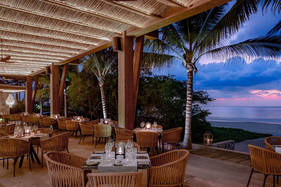 Beachfront restaurant pavilion with wooden dining tables, sunset ocean view, and palm trees