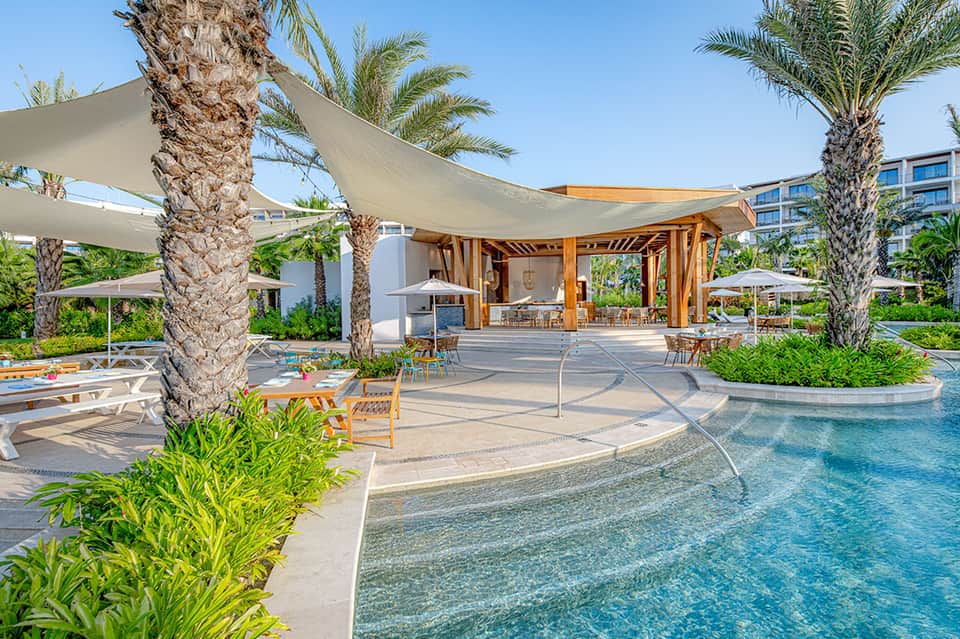 Luxury resort pool deck with palm trees, cabanas, sail shade structures, and lounging area