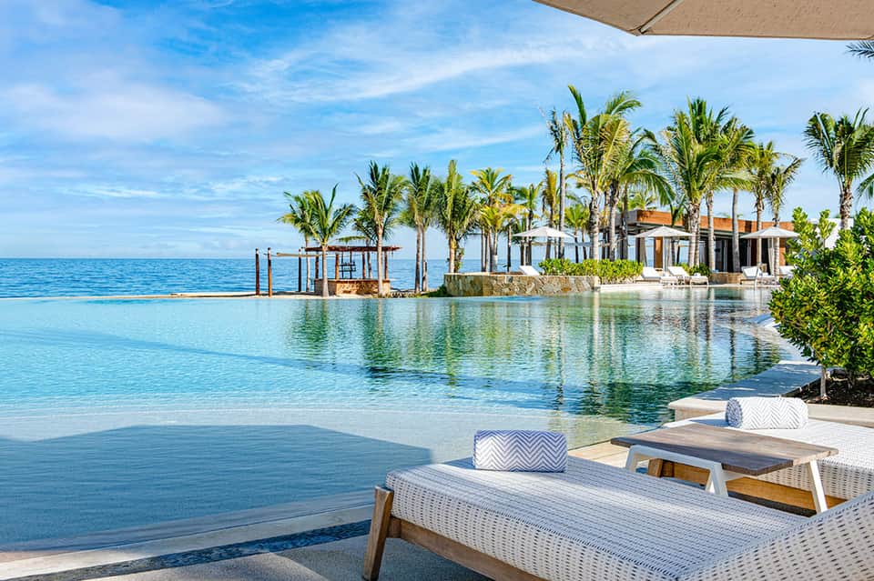 Luxury beachfront infinity pool with palm trees, turquoise water, white lounge chairs, and ocean horizon view