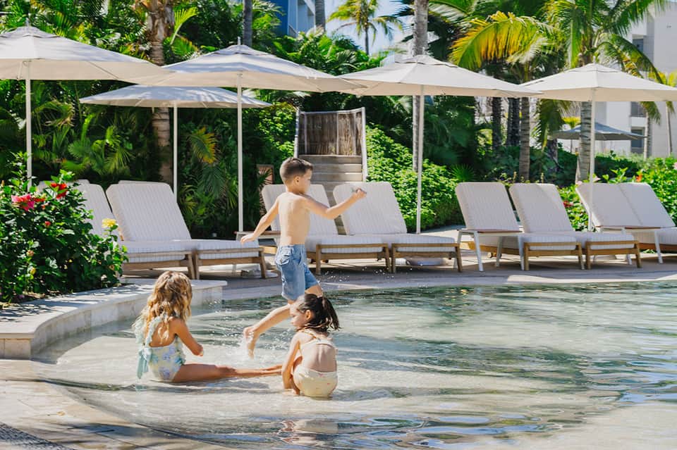 Shallow pool with children playing, white umbrellas, lounge chairs, and lush tropical vegetation