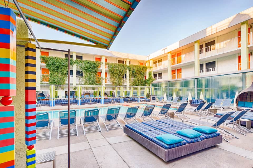 Colorful hotel courtyard with striped awning, lounge chairs, and modern residential buildings surrounding a sparkling pool.