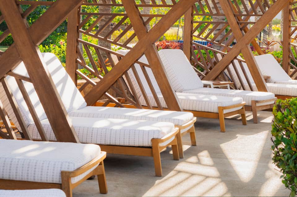 Wooden lounge chairs with white cushions arranged under a geometric wooden pergola in dappled sunlight
