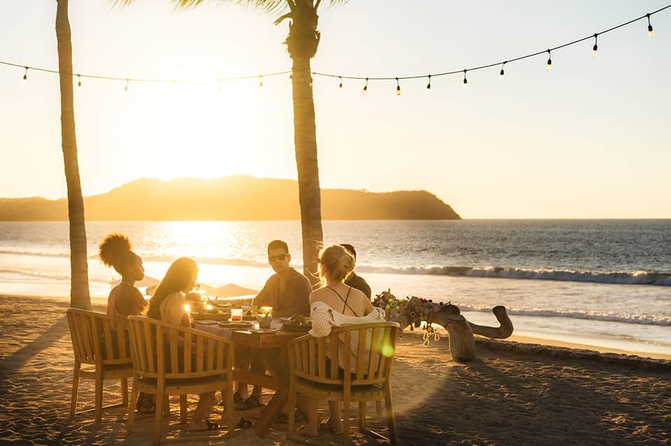 Group dining at beachfront table under string lights at sunset with ocean and palm trees