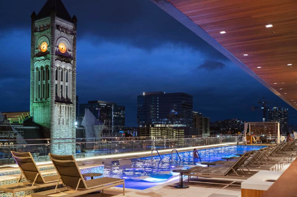 Illuminated rooftop pool at night with clock tower view, lounge chairs, and city lights backdrop