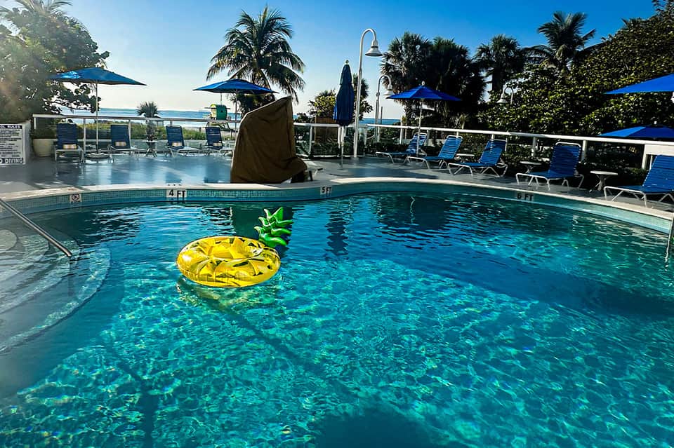 Resort pool with blue umbrellas, palm trees, pineapple-shaped pool float, and ocean view