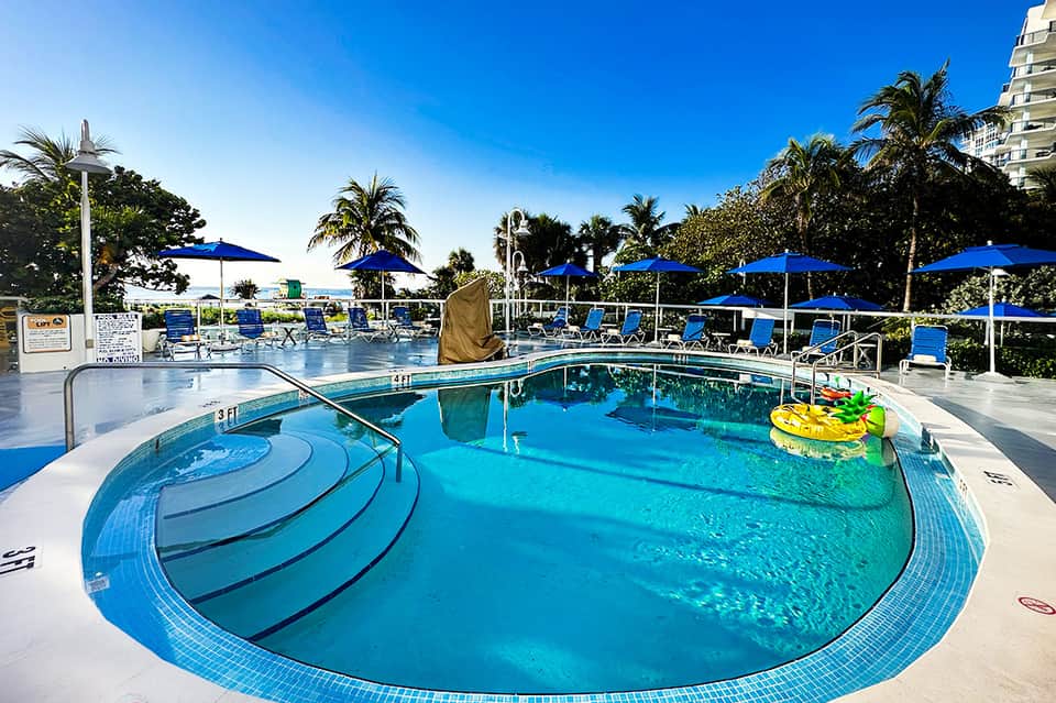 Resort pool with blue umbrellas, palm trees, lounge chairs, and coastal setting