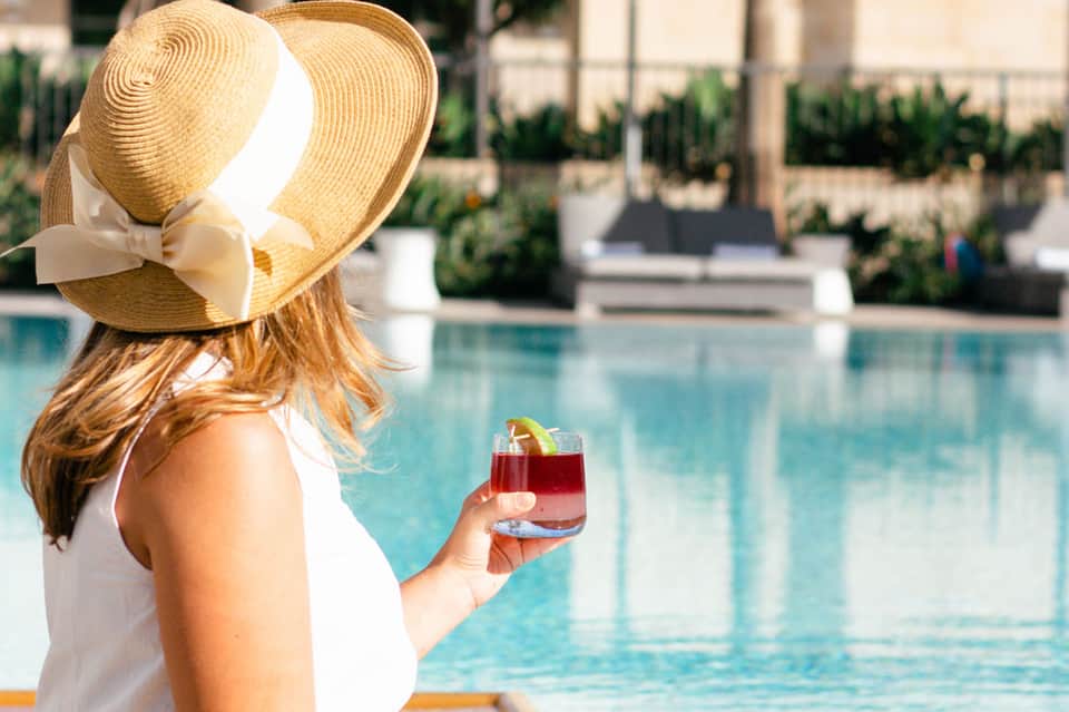 Woman in white swimsuit and straw hat holding tropical cocktail by resort pool with palm trees visible