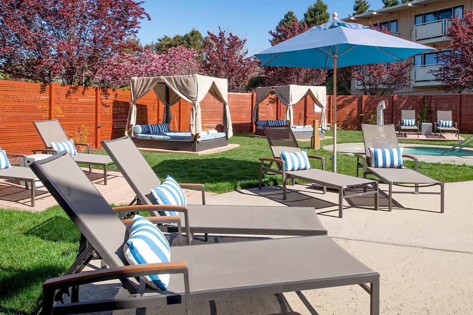 Resort pool area with white loungers, blue umbrellas, and enclosed cabanas surrounded by landscaping
