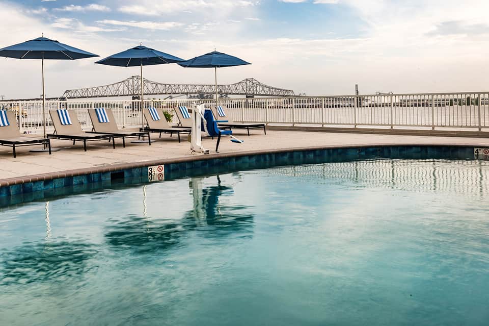 Outdoor pool with navy umbrellas, lounge chairs, and river bridge visible beyond white railing