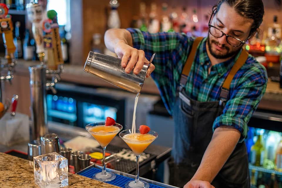 Bartender in plaid shirt pouring cocktail into martini glass at resort bar