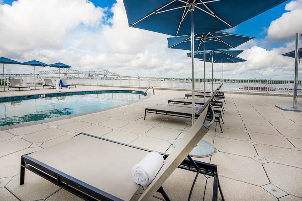 Rooftop pool with blue umbrellas, loungers, river view, and architectural bridge in background