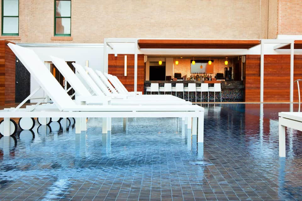 Modern hotel courtyard with reflective wet pavement, white lounge chairs, and indoor bar with warm lighting