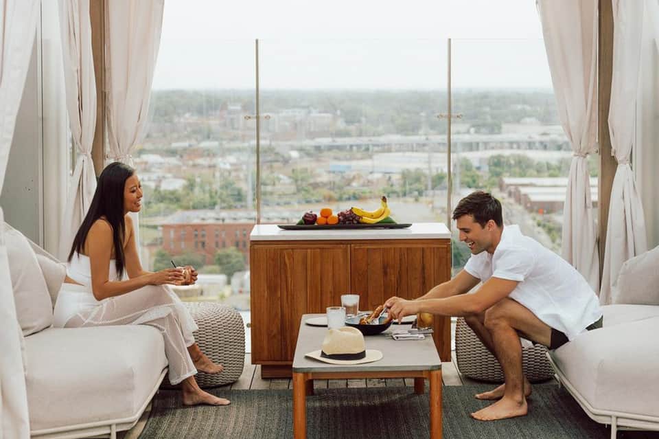 High-rise hotel room suite with two guests, floor-to-ceiling windows overlooking cityscape