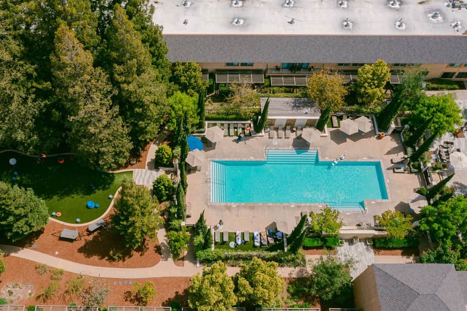 Aerial view of resort pool surrounded by trees, umbrellas, and manicured grounds with building in background