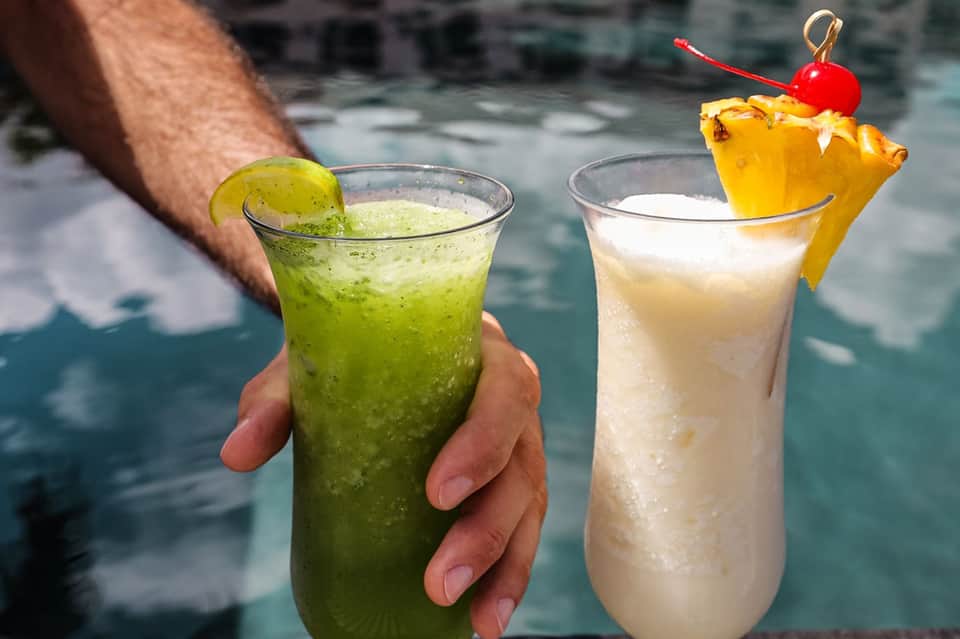 Two tropical cocktails held by person near pool, lime-colored and pineapple drinks
