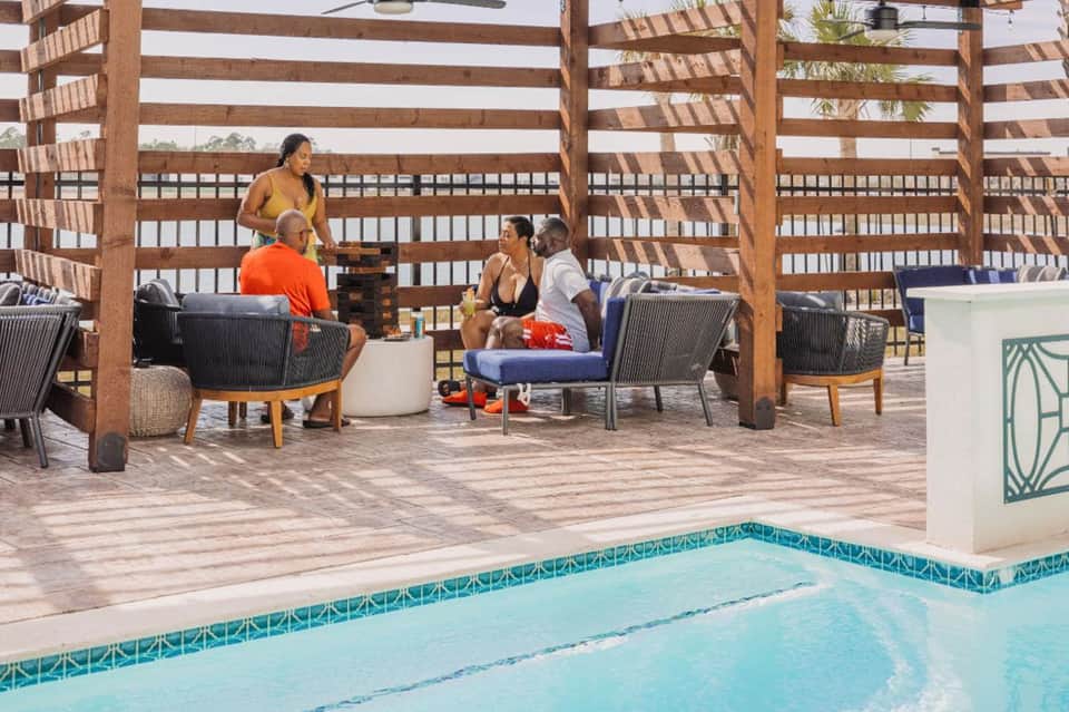 Resort guests relaxing in lounge chairs under striped wooden pergola by infinity pool and beach