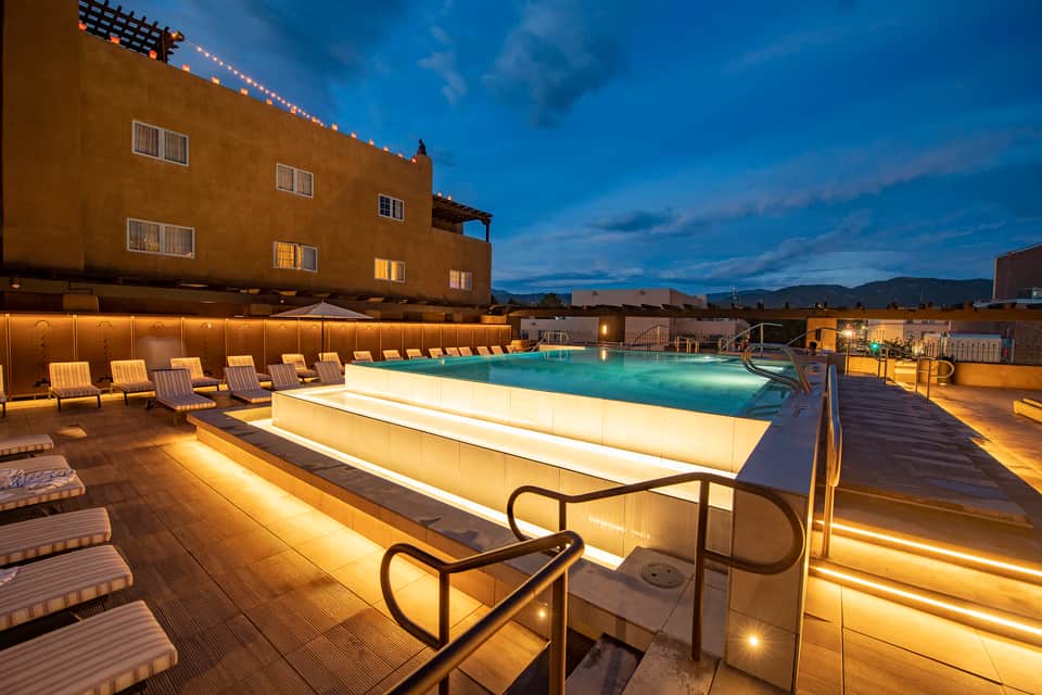 Illuminated rooftop pool at dusk with lounge chairs and mountain views