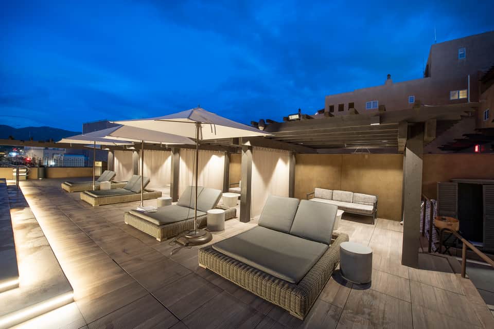 Illuminated rooftop terrace at dusk with woven loungers, cream umbrellas, cushioned seating, and ambient lighting