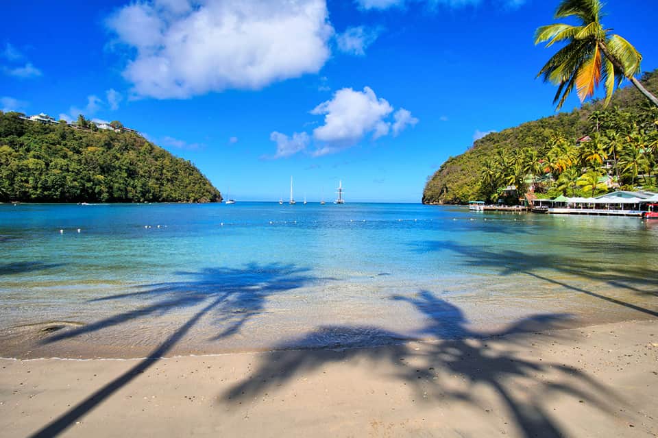 Tropical beach cove with clear turquoise water, palm tree shadows, and sailboats in distance